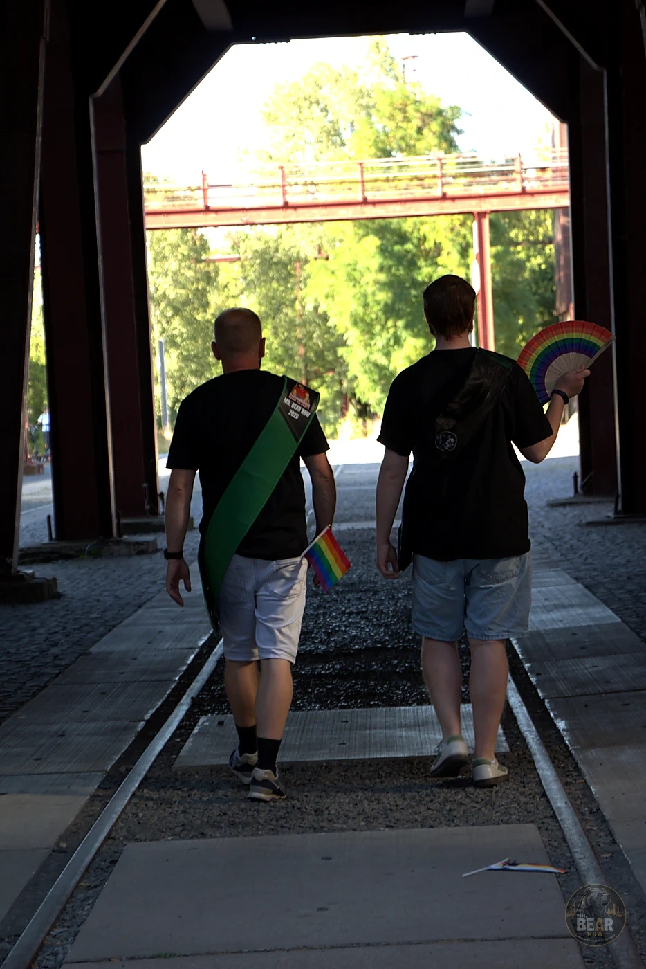 Christoph und Joshua gehen mit Pride-Fahne und Fächer durch die Industriekulisse