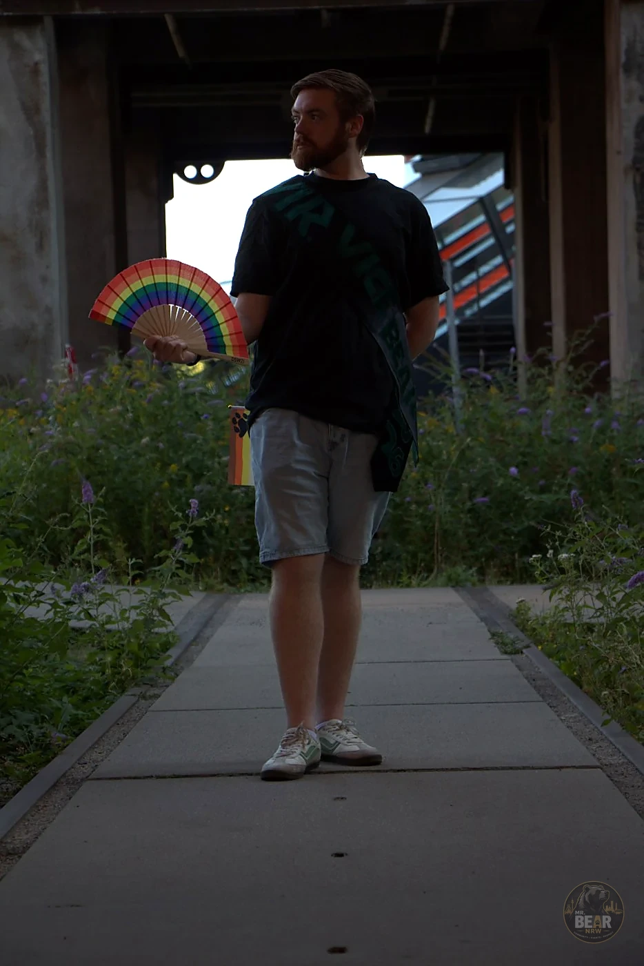Joshua mit Schärpe und Pride-Fächer zwischen Industriebauten